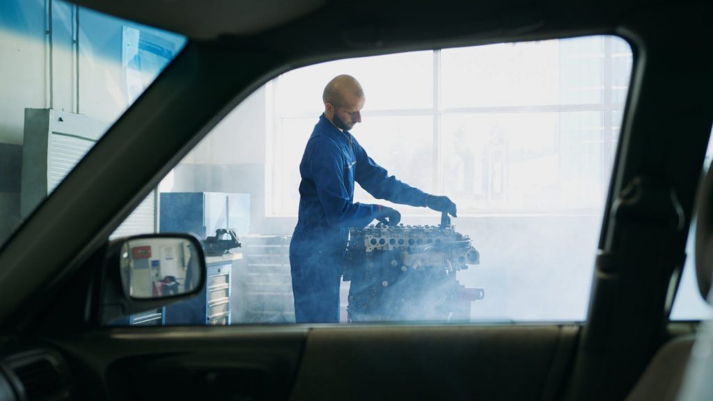 Mechanic in blue overalls working on an engine inside a garage with visible steam around.